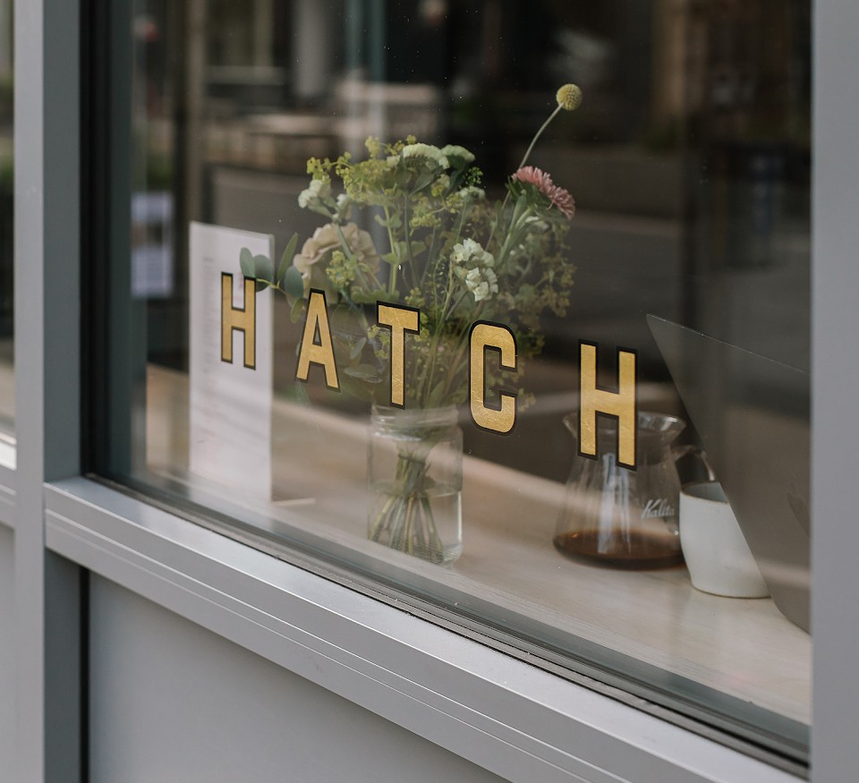 The Blackrock window: gold HATCH lettering on the glass, a jam jar of wildflowers and a Kalita Wave brewer on the counter, grey window frames, nobody in the shot.