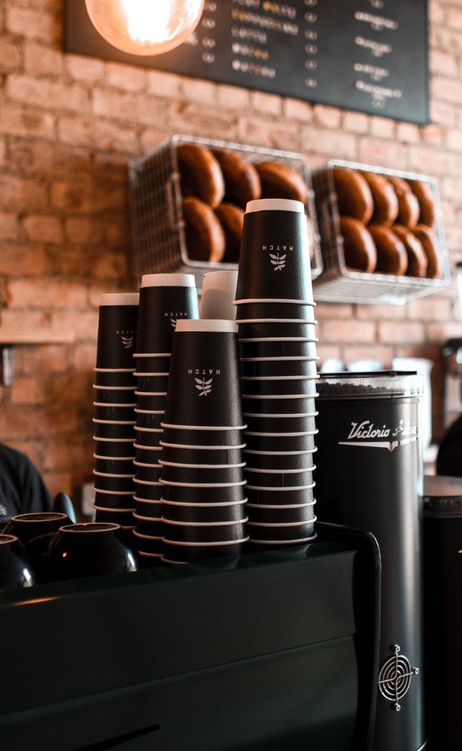 Stacked black cups printed with the sprig-and-HATCH mark, Victoria Arduino bean hoppers and a brick wall behind the counter at Glasthule.